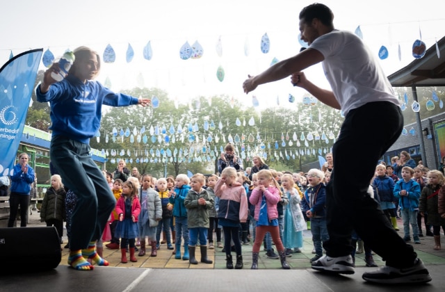 Acteur, danser en presentator Juvat Westendorp op een podium waarop een kind net een sprong maakt. De foto is genomen vanaf het podium, op de achtergrond staan tientallen basisschoolkinderen op een schoolplein met bioven hen slingers in de vorm van waterdruppels.