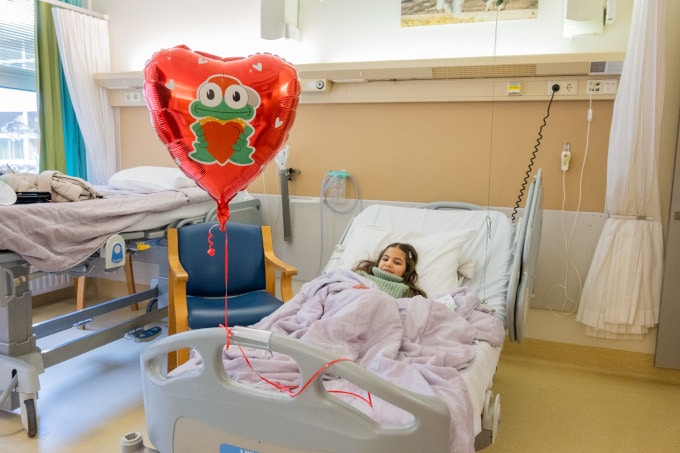 Meisje in ziekenhuisbed met aan haar bed een ballon in de vorm van een hart, waarop de kikker van Opkikker staat. De kikker heeft een rode pet op met de klep schuin naar achteren en heeft een hart in zijn handen.