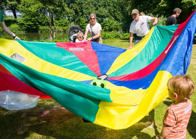 Een veelkleurige parachute met ongeveer in het midden een bal, die door enkele volwassenen en kinderen over de parachute wordt gerold. De parachute heeft blauw, geel, groen en rood als kleuren en de mensen staan in een parkachtige omgeving.